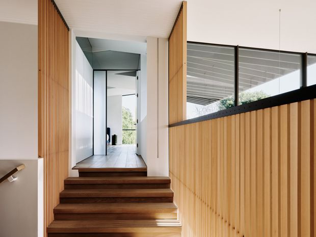 A bright contemporary interior hallway viewed from a timber staircase. The stair and adjacent walls feature warm vertical timber battens, while a black-framed glazed balustrade overlooks a lower level or adjacent space. Pale timber flooring, white walls, a white linear ceiling, and abundant natural light create a clean, refined transition space.
