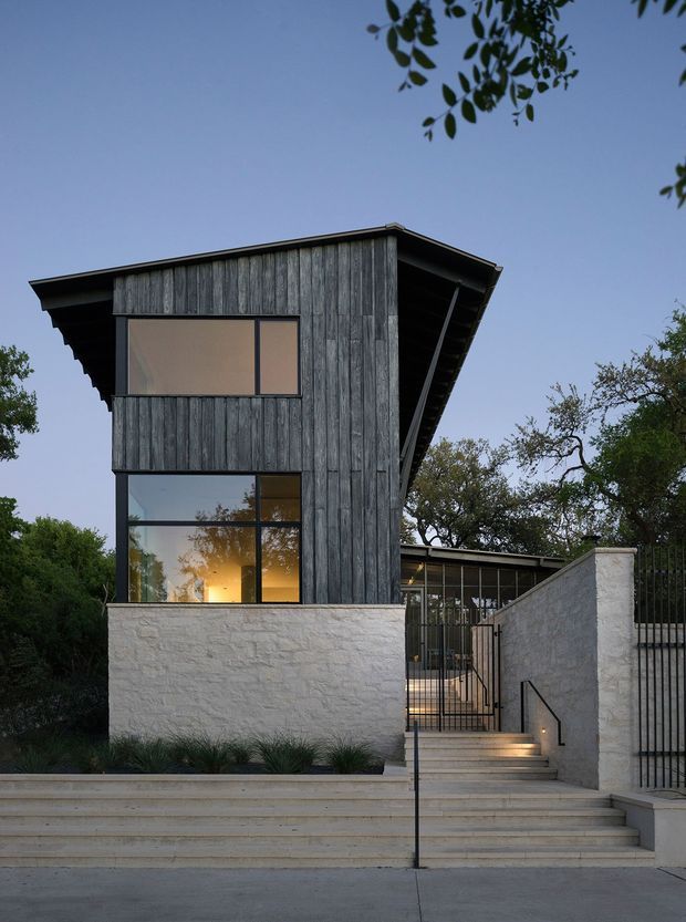 Exterior architectural photograph of a compact modern multi-level residence viewed from the street. The building features dark vertical timber cladding, large black-framed windows, a pronounced sloping roof with exposed eaves, pale masonry boundary walls, and a stepped entry path with black metal handrails and gates. Mature landscaping surrounds the home, with warm interior light visible through the lower window.