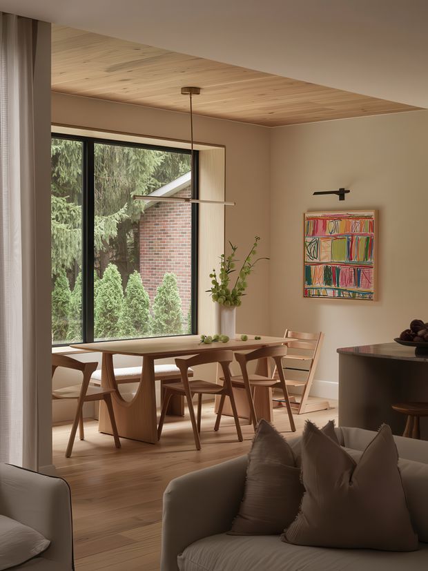 Warm, refined open-plan dining and living interior with a timber-lined ceiling, pale timber flooring, large black-framed picture window, and a sculptural timber dining table surrounded by mid-century-style chairs. A neutral upholstered sofa and cushions occupy the foreground, while a dark kitchen or sideboard counter appears at right. Soft beige walls, understated pendant and wall lighting, greenery, and colourful framed artwork create a calm, contemporary setting.