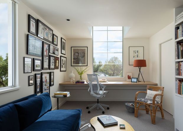 Bright, compact home office with a built-in desk spanning beneath a large window and skylight. The room includes a task chair, extensive framed artwork and photographs on one wall, a bookcase, a blue sofa, a rattan armchair, and a small round coffee table. Neutral walls and carpet are balanced by warm timber finishes and blue upholstery.