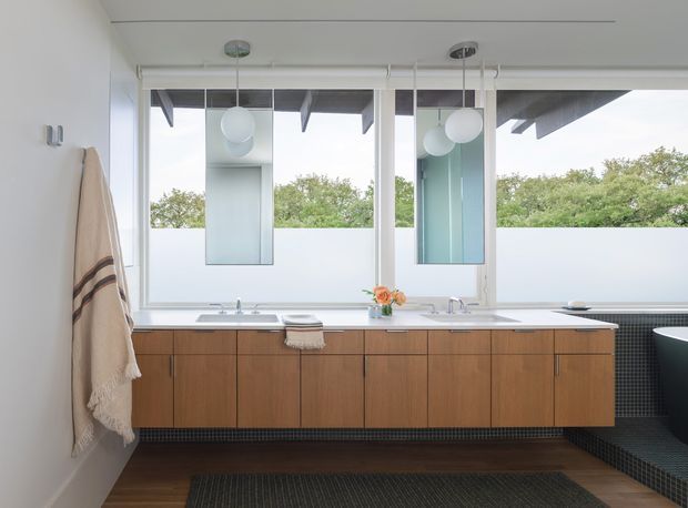 Bright, mid-century-inspired bathroom vanity beneath a wide bank of windows. The room features a long timber vanity with integrated basins and chrome taps, white benchtop, dark small-format tiled wall and floor accents, suspended white globe pendant lights, mirrored wall cabinets, and a partially visible freestanding bathtub at right. The windows provide abundant natural light and views of greenery.