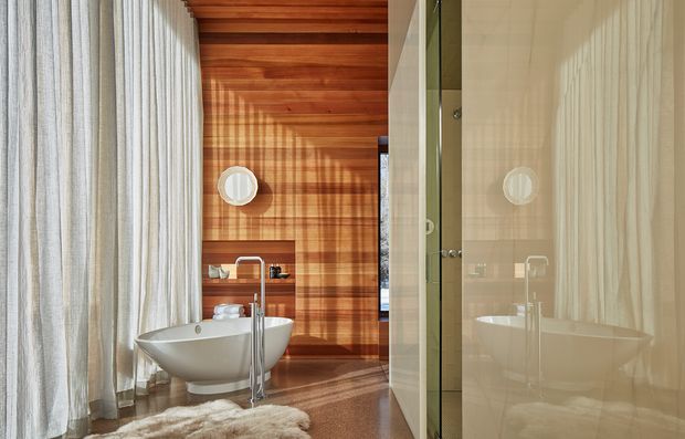 A refined bathroom with a freestanding white oval bathtub positioned beside a tall brushed-metal floor-mounted tap. The room features warm timber wall and ceiling lining, a large glass shower enclosure, sheer full-height curtains, and a pale glossy tiled or stone wall reflected in the glazing. Soft daylight creates strong linear shadows across the timber, while a textured cream rug adds warmth.
