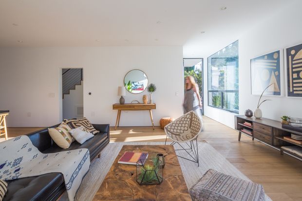Bright contemporary living room with white walls, light timber flooring, expansive floor-to-ceiling glazing, and a mix of mid-century-inspired furniture and patterned textiles. The room includes a dark leather sofa, patterned armchair, substantial timber coffee table, console table with round mirror, and abstract wall art. Natural light creates an airy, warm, inviting atmosphere.