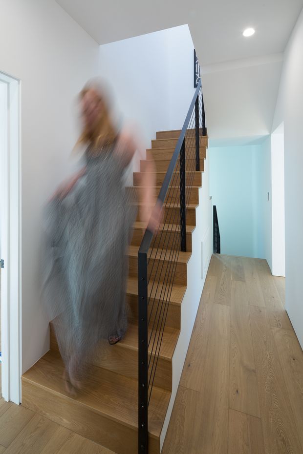 Bright contemporary interior hallway focused on a straight timber staircase with slim black metal railing and vertical balusters. The staircase rises beside a white wall, with a pale timber floor continuing through the corridor and a light blue accent wall visible beyond. A blurred person is moving on the stairs, creating a sense of motion. The space is clean, minimal, and naturally bright, with recessed ceiling lighting.