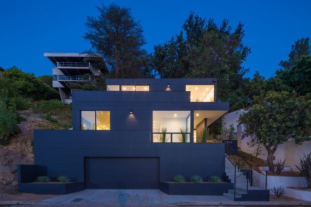 Twilight street-facing view of a contemporary multi-level residence with a dark, boxy facade, flat rooflines, broad rectangular glazing, recessed lighting, an integrated garage, planted raised beds, and an external stairway. A neighboring or upper-level structure is visible behind the main house, with mature landscaping surrounding the property.