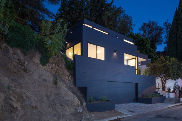 Twilight exterior view of a modern, flat-roofed multi-level residence built into a steep sloped site. The dark horizontal-panel facade features large illuminated windows, narrow clerestory glazing, a projecting upper volume, and an integrated garage at street level. A paved entry walkway and planted terraces lead up beside the house, with mature trees surrounding the property.