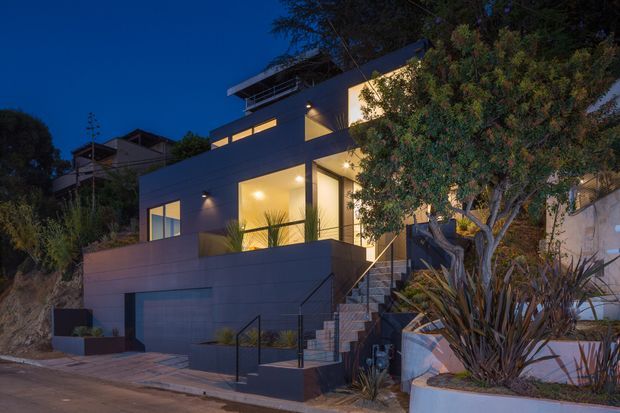 Twilight exterior view of a contemporary multi-level home on a sloped site. The house has clean horizontal lines, dark rendered or clad walls, extensive illuminated glazing, roof terraces or projecting flat roof planes, an integrated garage, and a stepped entry stair. Mature landscaping and raised planters frame the facade.