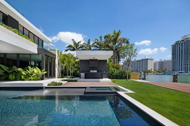 A contemporary waterfront residential exterior with a geometric swimming pool in the foreground, a manicured lawn and timber deck beside the water, and a covered outdoor entertaining pavilion with a built-in fireplace or barbecue feature. The house has crisp white rendered forms, expansive dark-framed glazing, glass balustrades, and lush tropical planting. High-rise waterfront buildings are visible across the water.