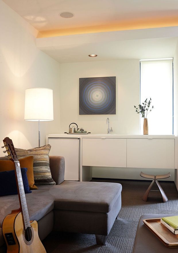 A compact, refined living room with a large grey sectional sofa, guitar, floor lamp, patterned cushions, and a low coffee table. Along the rear wall is a minimalist white built-in cabinet or console with a small sink-like fitting, artwork above, and a narrow window with a roller blind. Recessed ceiling lighting and warm concealed cove lighting create a comfortable contemporary atmosphere.