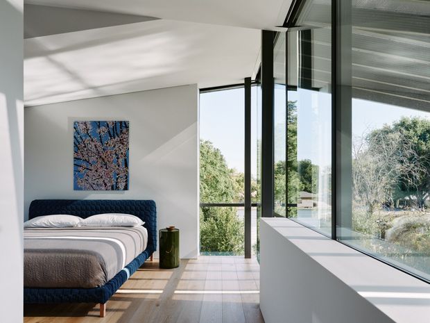 Bright contemporary bedroom with a low-profile upholstered navy bed, layered neutral bedding, white walls, pale timber flooring, and extensive floor-to-ceiling glazing. A long white built-in window seat or ledge runs along the windows, which overlook landscaped greenery. Strong sunlight creates geometric shadows across the floor and walls, giving the room an airy, refined feel.