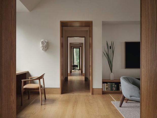 A warm, refined interior view looking through a sequence of timber-trimmed doorways into a living area and further rooms. The foreground has a timber floor, a compact writing desk with a wooden chair, and a sculptural wall ornament. At right is part of a living room with a grey upholstered armchair, low timber media cabinetry, a wall-mounted television, and a tall decorative plant. Natural timber framing and restrained neutral finishes create a calm, cohesive composition.