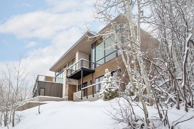 Exterior view of a contemporary multi-level house set on a snowy, wooded site. The building has a strongly angular form, broad roof overhangs, warm-toned horizontal cladding, dark framed glazing, and multiple projecting balconies and terraces with black metal railings. Snow-covered ground and deciduous trees surround the residence.