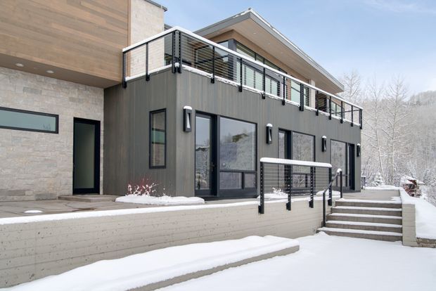 Exterior view of a contemporary multi-level house in a snowy setting. The building combines dark vertical cladding, pale stone wall sections, expansive black-framed glazing, and open metal balustrades. Broad stepped terraces and retaining walls lead through the snow-covered outdoor area, while a projecting upper volume creates a sheltered balcony.