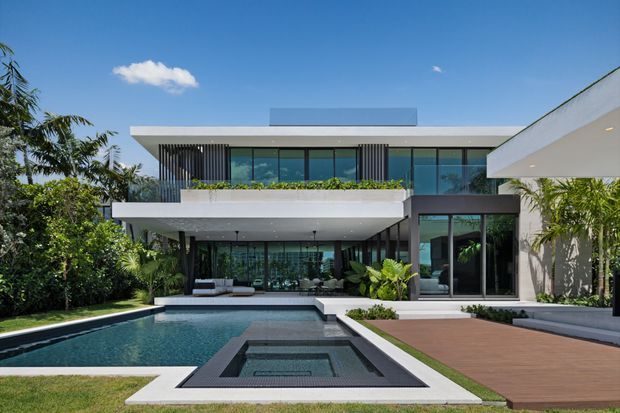 A contemporary multi-level residence viewed from the landscaped pool area. The home features strong horizontal flat roof slabs, extensive floor-to-ceiling glazing, dark vertical facade screens, white rendered surfaces, a planted upper terrace, and a timber-toned deck surrounding a swimming pool and square spa.