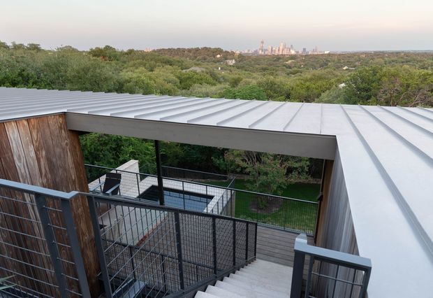 Elevated exterior view of a modern multi-level home with a broad corrugated metal roof, weathered timber wall cladding, dark metal railings, and an outdoor stair and deck system overlooking dense greenery and a distant urban skyline.