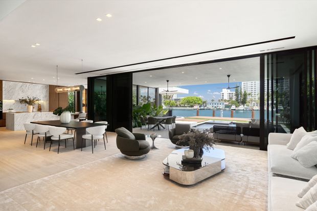 A bright, spacious open-plan living and dining area with full-height retractable glazing opening toward a waterfront view. The interior features pale timber flooring, a large light neutral rug, a white sectional sofa, sculptural lounge chairs, a low stone or concrete coffee table, a dining table with curved upholstered chairs, a marble feature wall, and dark-framed glazing and joinery. The room has a refined contemporary aesthetic and strong indoor-outdoor connection.
