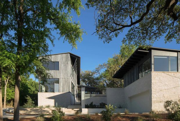 Exterior view of a contemporary low-rise residential property composed of two angular pavilion-like volumes around a landscaped entry courtyard. The buildings feature flat or very low-pitched roof forms, expansive rectangular glazing, pale textured masonry walls, and vertical timber-like cladding, set among mature trees under a clear blue sky.