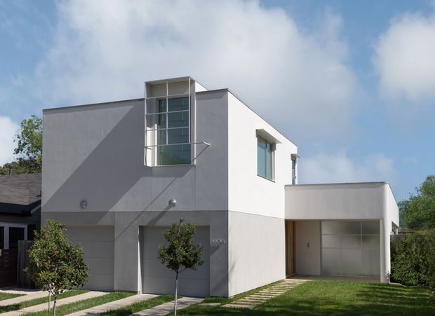 Exterior photograph of a contemporary two-storey white and light-grey rendered home. The facade has a cubic, flat-roofed composition, a tall glazed stair or double-height window, simple rectangular windows, an attached single-storey garage, and a paved path through a landscaped front lawn.