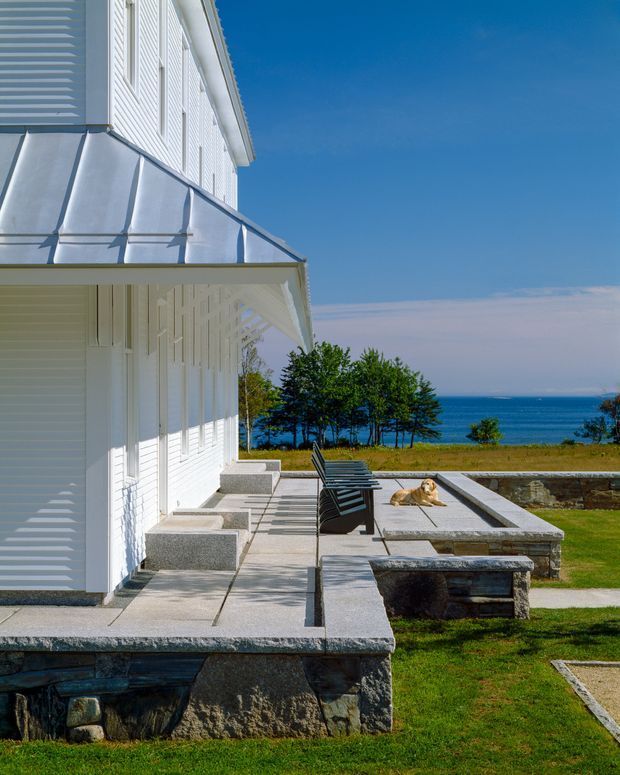 Exterior architectural photograph of a crisp white coastal-style house with horizontal siding, a standing-seam metal roof and broad covered terrace. Stone retaining walls and paved outdoor seating terraces step down toward a bright blue body of water, with lawn and trees surrounding the property. The composition emphasizes the building facade, roof details, and waterfront outdoor setting under a clear sky.