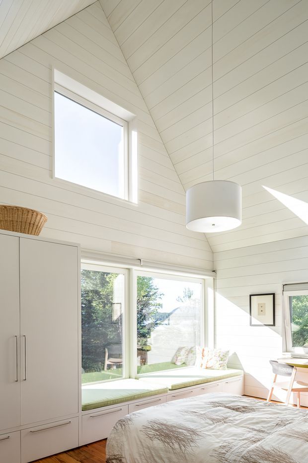 Bright, airy bedroom with a steeply vaulted ceiling clad in white-painted horizontal boards. The room features built-in white cabinetry, a window-seat bench with pale green cushions, large windows, a simple white pendant light, timber flooring, and a partially visible bed. Sunlight creates strong highlights across the space.