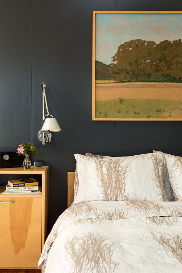 A warmly styled bedroom vignette with a dark charcoal wall, light timber bedside cabinet, white and taupe botanical-patterned bedding, a wall-mounted swing-arm lamp, small decor, and a framed landscape artwork in a timber frame. The composition feels refined, cosy, and slightly mid-century inspired.