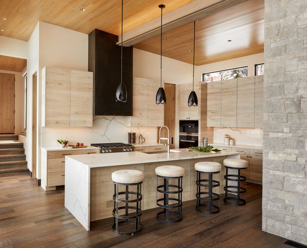 A spacious, open-plan kitchen with a large marble-look island and seating for four. The room features light timber cabinetry, dark wood flooring, a warm timber ceiling, a dark feature rangehood, black pendant lights, integrated appliances, and a substantial stacked-stone wall. Steps and an adjoining circulation area are visible to the left.