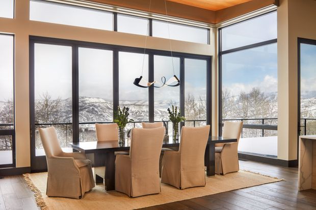 A bright, refined dining area with a dark rectangular dining table and six upholstered slipcovered chairs, positioned beside expansive corner glazing. Snow-covered mountains and trees are visible beyond the windows. The room has warm timber ceiling detailing, dark window frames, light neutral walls, a pale area rug, and a contemporary suspended sculptural light.
