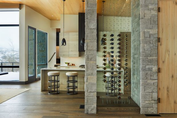 Open-plan contemporary interior featuring a stone-clad structural column and wall framing a glass-enclosed wine display, with a compact kitchen island and two bar stools beyond. The room has warm timber flooring and a timber-lined ceiling, large glazed doors, dark-framed pendant lights, and integrated timber cabinetry.