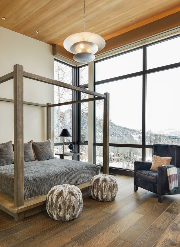 A warm, contemporary bedroom with a substantial four-poster timber bed, textured grey bedding, and sculptural round upholstered stools. Floor-to-ceiling black-framed glazing overlooks a snowy, wooded mountain landscape. The room features a timber-clad ceiling, wide-plank timber flooring, a dark leather armchair, and a layered metallic pendant light.