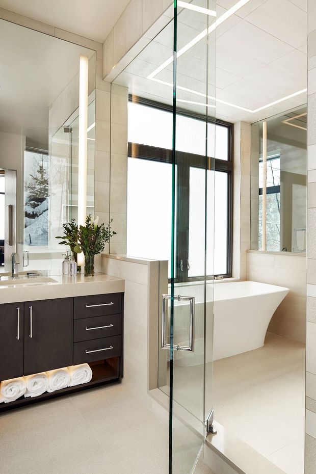 A bright, contemporary bathroom with a large frameless glass shower enclosure, freestanding white bathtub, dark floating vanity with integrated basin, expansive mirrors, and a tall black-framed window. Pale large-format tiled walls and floor create a clean, light-filled setting, with linear recessed ceiling lighting and minimal decorative styling.