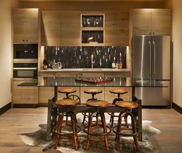 A warm, moody contemporary kitchen with extensive wood cabinetry, integrated appliances, a stainless-steel refrigerator, and a dramatic dark stone or patterned splashback. A central island with a dark framed base and timber top is paired with multiple vintage-style wooden bar stools, set over a hide rug on timber flooring. The kitchen includes open display shelving, under-cabinet lighting, and a compact dining or entertaining layout.
