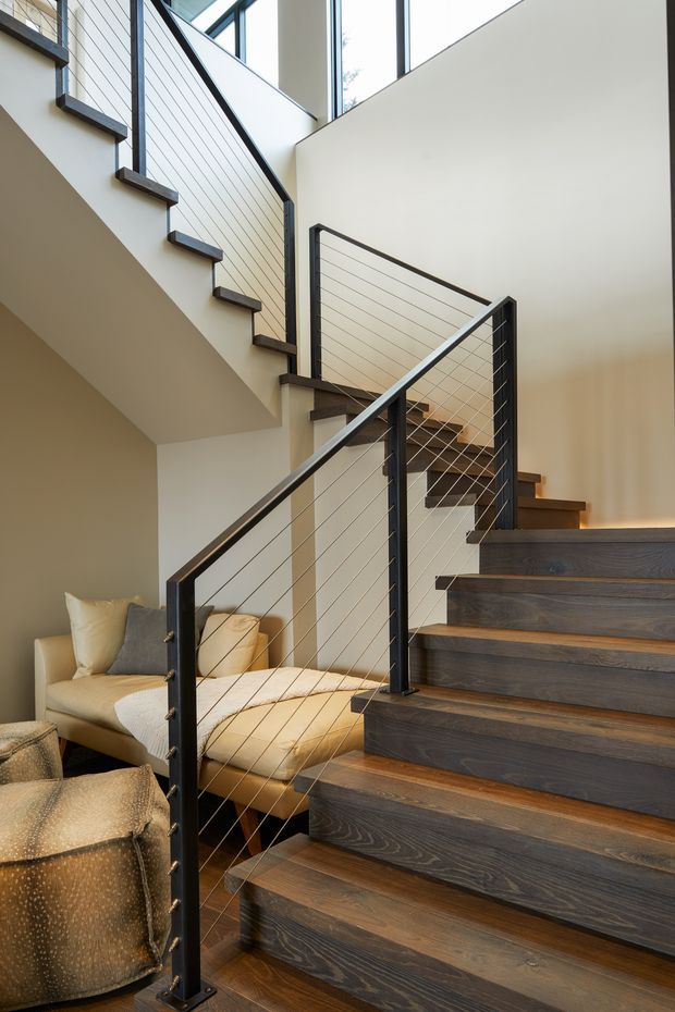 Interior photograph looking up a modern staircase with dark timber treads, slim black metal posts and horizontal cable balustrades. The stair rises through a bright, open void with tall glazing and pale walls, while a comfortable seating area is visible beneath and beside the stairs. The space has warm layered lighting and a contemporary, clean-lined design.