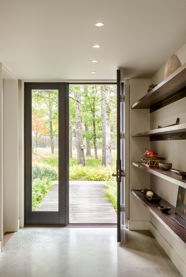 A bright, minimalist entryway opening onto a timber deck and leafy garden. The interior features a smooth pale floor, warm off-white walls, recessed ceiling lights, a dark-framed glazed door, and built-in open display shelving with dark timber shelves along the right wall.