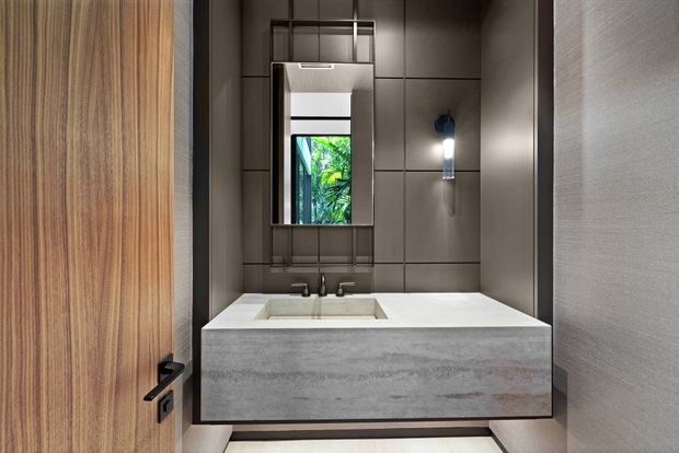 Compact, high-end bathroom vanity viewed from an open timber door. The room features a floating grey concrete-look vanity with an integrated rectangular basin and dual tapware, dark large-format wall panels, a narrow vertical mirror with metal framing, a contemporary wall sconce, and a small window or reflected view of greenery. The design is restrained, contemporary, and strongly material-focused.