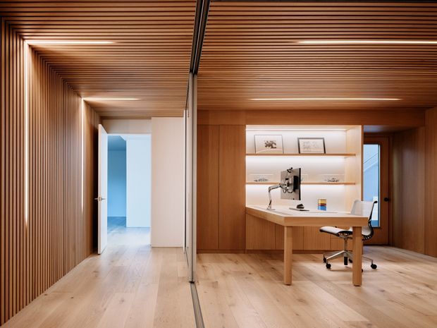 A refined contemporary home office with a built-in timber desk and illuminated open shelving, viewed through a full-height glazed partition. The room features pale timber flooring, warm timber wall and ceiling lining, integrated linear lighting, and a black ergonomic office chair. A ribbed timber-lined corridor and an adjacent bright blue-accented room are visible beyond the office.