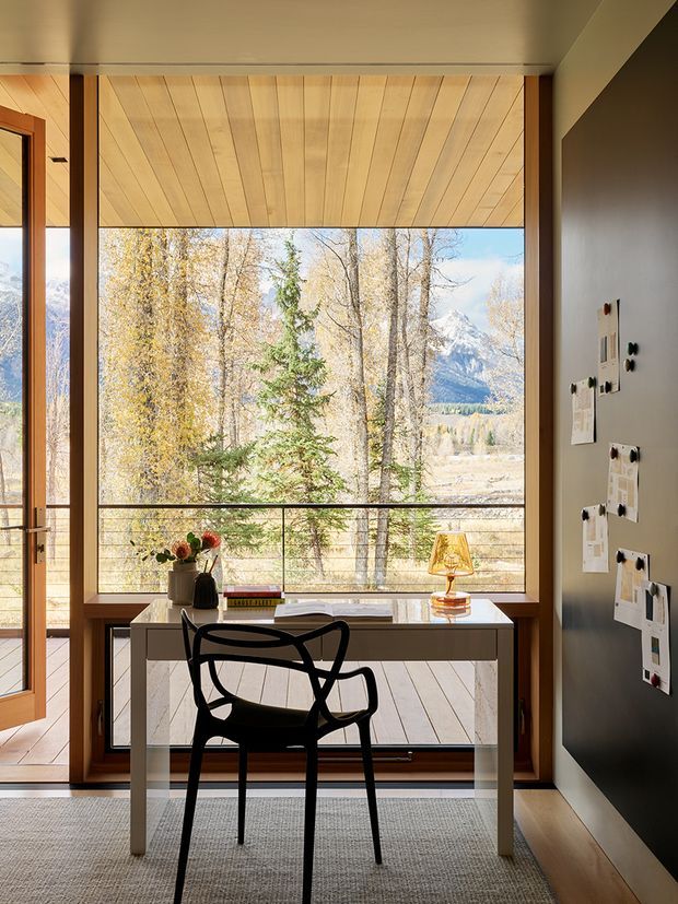 A compact, refined home office positioned in front of expansive glazing with a mountain and forest view. The room features a simple white desk, black sculptural chair, small table lamp, flowers, wall-mounted notes, timber-framed glazing, warm timber ceiling panels, dark painted wall, and a neutral rug. The space is bright and visually connected to the alpine landscape.