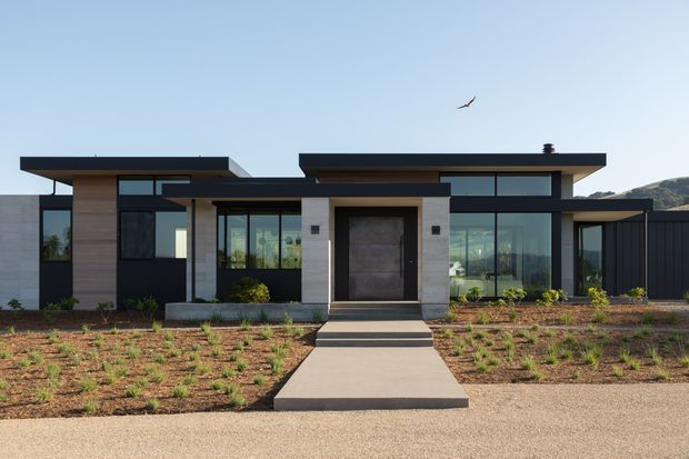 Front exterior view of a contemporary single-storey residence with a strong horizontal composition, flat roof planes, extensive dark-framed glazing, timber-look wall cladding, pale masonry or concrete portal elements, and a central recessed entry. A straight concrete path crosses a sparsely planted landscaped forecourt.