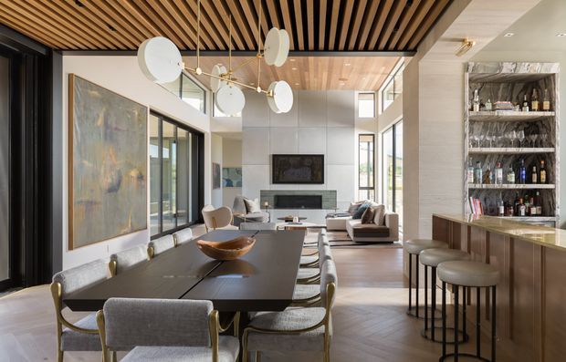A sophisticated open-plan living and dining space with a built-in home bar. The room features a long dark dining table with upholstered dining chairs, a contemporary lounge area, a fireplace and television wall, expansive black-framed glazing, and a dramatic slatted timber ceiling. A sculptural multi-disc pendant light hangs above the dining area, while a marble-toned bar and glass-fronted bottle shelving occupy the right side. The palette is warm and neutral, combining timber, dark metal, pale stone-like surfaces, and soft grey upholstery.
