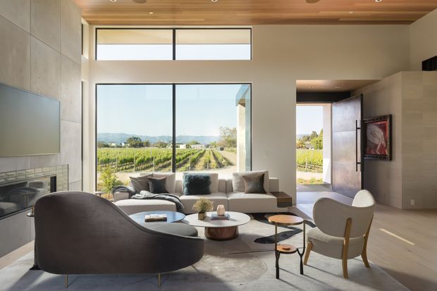 Bright contemporary living room with expansive floor-to-ceiling glazing overlooking vineyard rows. The room features a large sectional sofa, sculptural lounge chairs, round coffee tables, a linear fireplace with a wall-mounted television, a warm timber ceiling, pale stone or plaster walls, and an open doorway to the exterior.