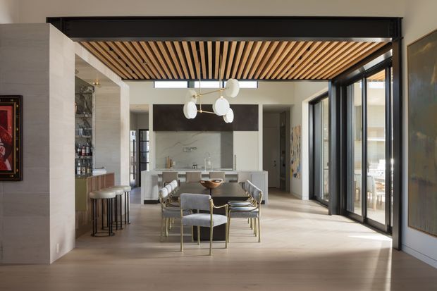 A refined open-plan interior viewed from a living or circulation area, looking toward a dining space and kitchen. The room features pale timber flooring, a slatted timber ceiling with dark framing, full-height black-framed glazing and doors, a marble or pale stone kitchen backsplash, and contemporary dining furniture beneath a sculptural pendant light. A bar area with stools and bottle storage is visible to the left, while bright outdoor space is visible through the glazing on the right.