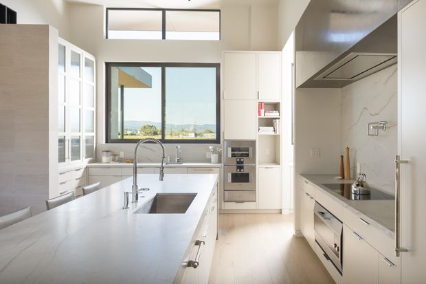 Bright contemporary galley-style kitchen with a large central island, integrated sink and brushed metal tapware, white custom cabinetry, pale timber flooring, and a marble-look stone splashback and benchtop. Floor-to-ceiling cabinetry and clerestory glazing create an airy, spacious feel, with views of a distant landscape through the rear window.