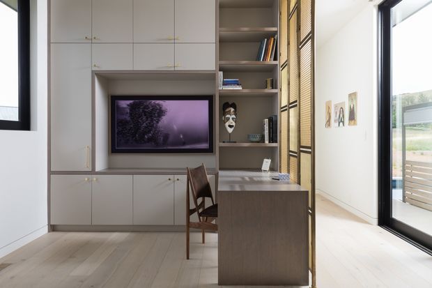 A bright, compact multifunctional interior featuring a full-height built-in storage wall with concealed cabinetry, open shelving, and an integrated television. A small timber desk projects from the joinery beside a simple wooden chair. A perforated or slatted timber screen separates the workspace from a light-filled area with large black-framed glazing and an outdoor deck visible beyond. Pale timber flooring and white walls create a clean, contemporary atmosphere.