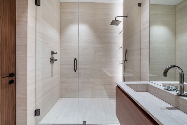 A contemporary bathroom featuring a large frameless glass walk-in shower, pale beige stone-look wall and floor tiles, and a timber-toned floating vanity with a light stone benchtop and integrated basin. Chrome fixtures, a curved tap, and a mirrored wall create a clean, refined appearance.