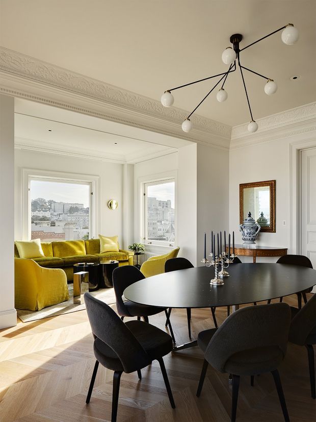 Elegant, light-filled open-plan dining and living space with a large oval black dining table, upholstered dark dining chairs, and a sculptural branching pendant light. The adjoining sitting area contains bright mustard-yellow sofas and armchairs beside tall bay windows overlooking an urban cityscape. Ornate white ceiling cornices, decorative wall mouldings, herringbone timber flooring, and classic trim are paired with contemporary furniture and lighting.