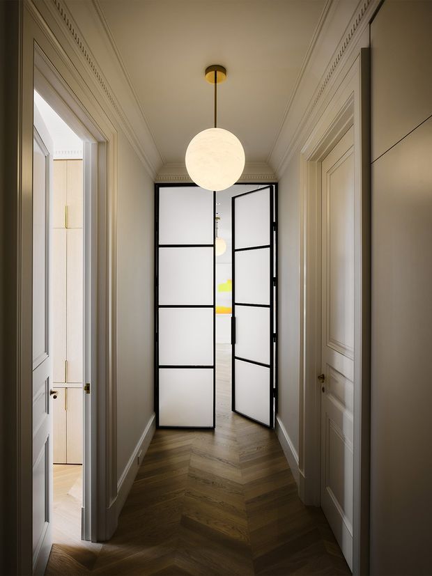 A narrow, refined residential hallway with warm herringbone timber flooring, cream-painted decorative wall panelling and ornate cornices. White paneled doors line both sides, while black-framed frosted-glass double doors lead to a brighter adjoining room. A spherical pendant light hangs centrally from the ceiling, creating a softly lit, elegant corridor.