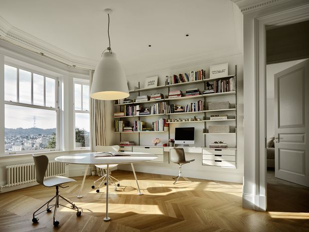 Bright, refined home office with a large bay window overlooking a cityscape, built-in wall-to-wall shelving and desk joinery, a round white work table, ergonomic chairs, and a sculptural pendant light. The room features ornate ceiling cornices, pale walls, white cabinetry, and a warm herringbone timber floor, with a doorway opening to an adjacent room.