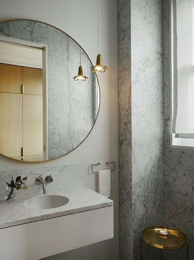 A refined, compact bathroom with a floating white vanity, integrated oval stone basin, wall-mounted chrome tapware, and extensive pale veined marble wall and benchtop surfaces. A large round mirror with a slim brass frame reflects warm timber cabinetry and decorative brass pendant lights. A small gold-toned stool and white towel add understated luxury, while a roller blind filters daylight through the window.