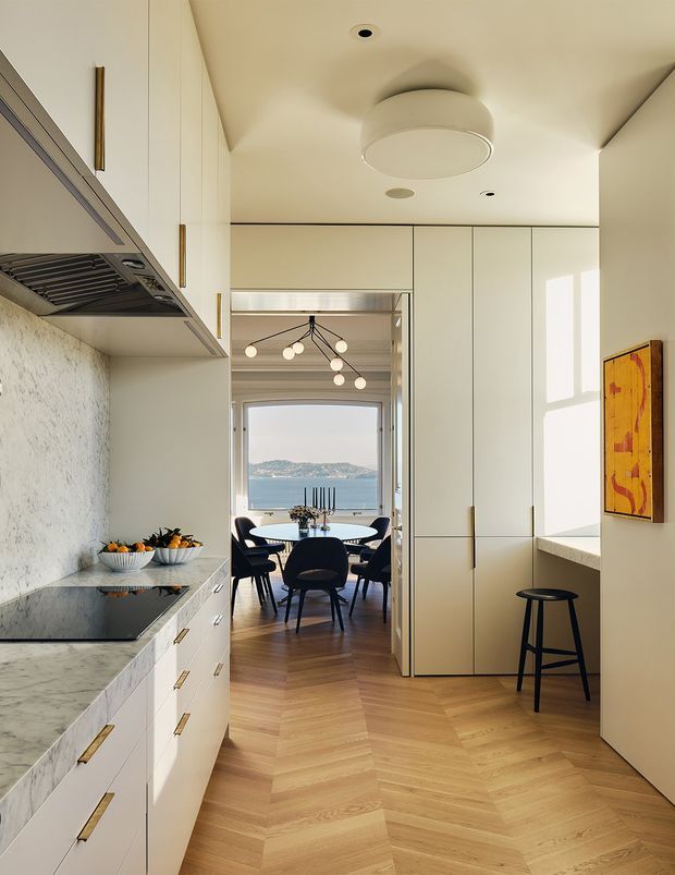 A warm, refined contemporary kitchen viewed toward an adjoining dining area and waterfront view. The kitchen features full-height pale cabinetry with slim brass-toned pulls, a marble-look stone benchtop and splashback, integrated appliances, and a herringbone timber floor. A large central opening leads to a dining room with dark chairs and a sculptural pendant light; sunlight casts strong rectangular shadows across the cabinetry and wall.