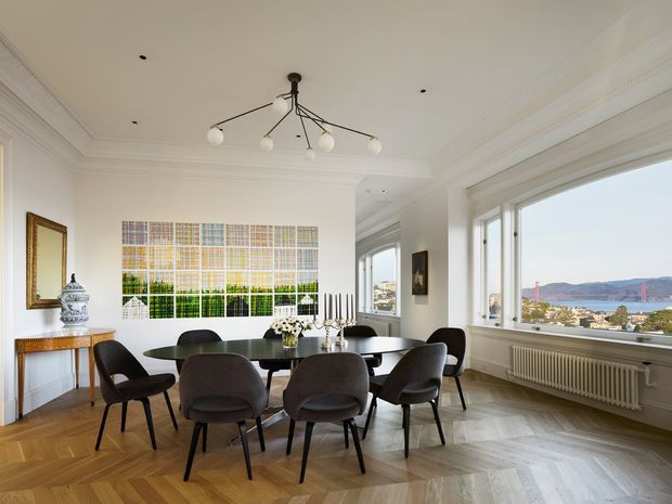 Elegant, light-filled dining room with a large dark dining table surrounded by upholstered chairs, a sculptural multi-arm pendant, white walls with ornate cornices, and a herringbone timber floor. Large windows overlook a coastal or waterfront landscape, while a decorative console, mirror, artwork, and radiator complete the refined interior.
