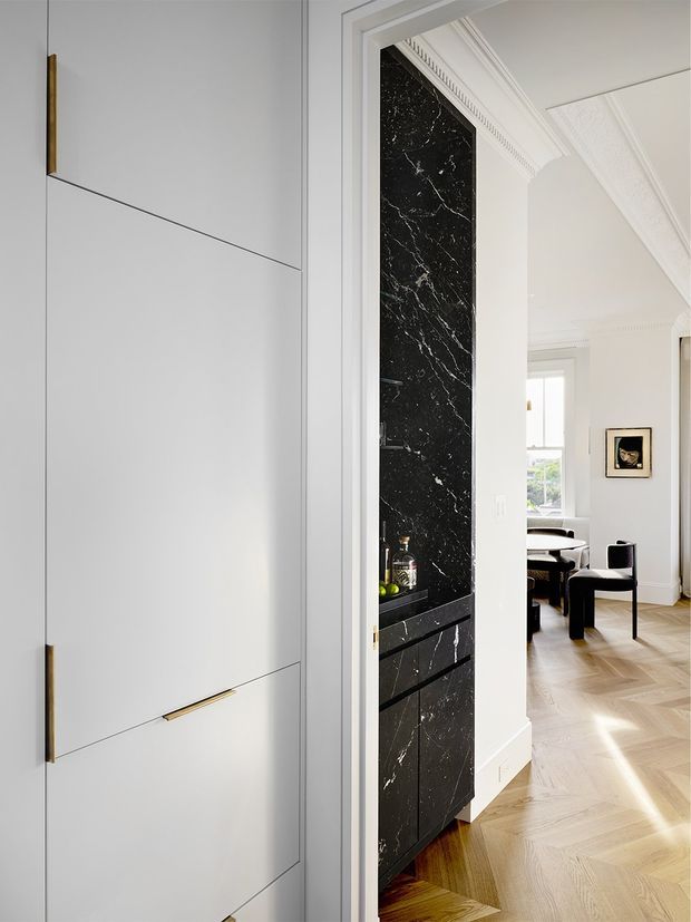 A bright, refined interior view showing a concealed white built-in cabinet with slim brass pulls in the foreground, beside a striking black marble or stone feature wall with integrated dark cabinetry and open shelving. Through the doorway is an elegant dining area with black furniture, herringbone timber flooring, white walls, tall windows, and ornate ceiling cornices and architraves.